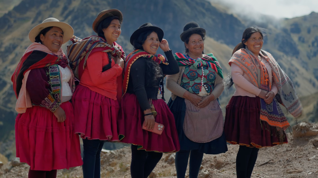 Five Andean women in colorful traditional outfits posing on a mountain trail, smiling.